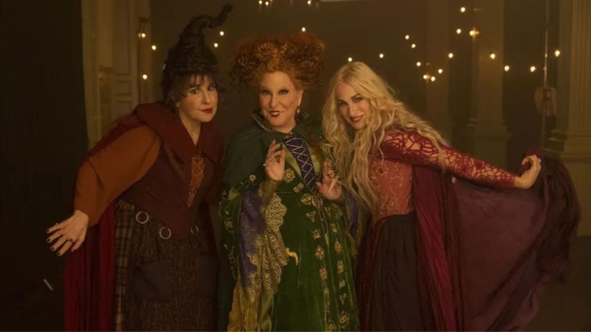 A imagem mostra as atrizes Bette Midler, Sarah Jessica Parker e Kathy Najimy fantasiadas de bruxas com vestidos volumosos e capas para interpretar as irmãs de Salem em Abracadabra 2.