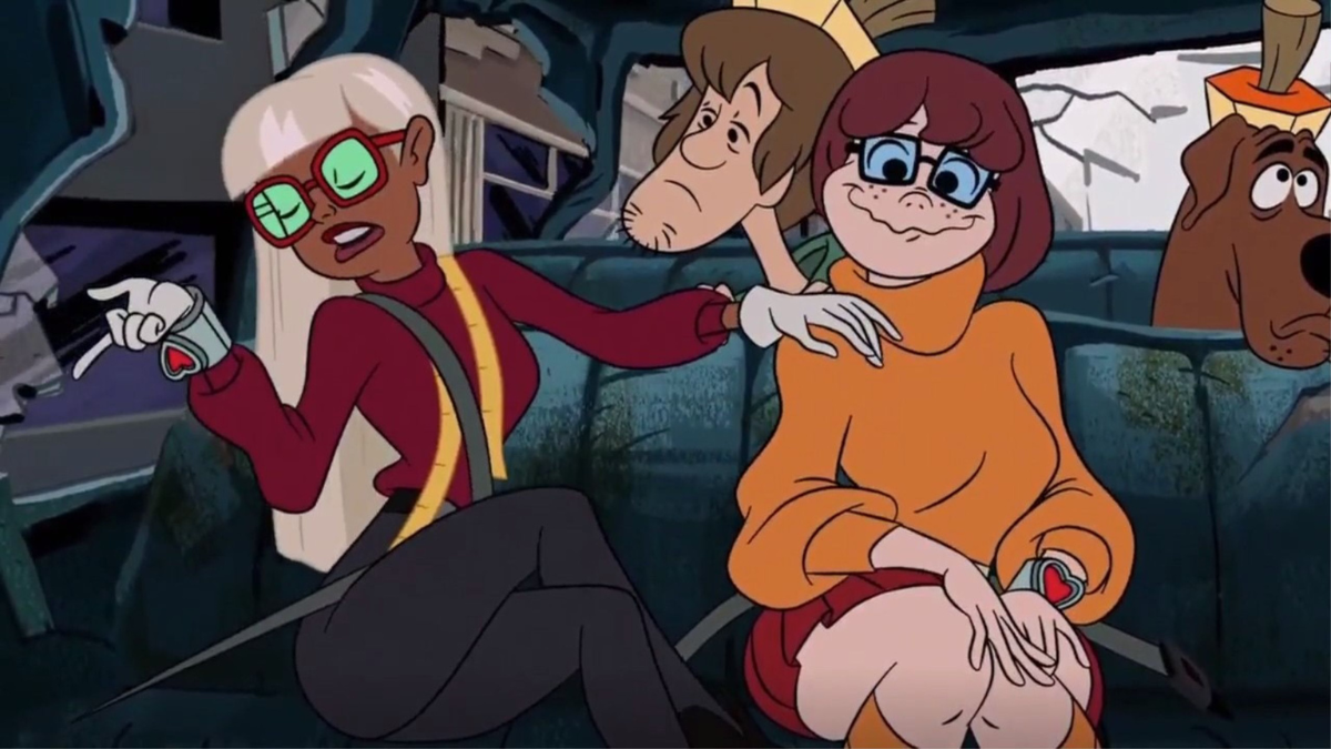 A imagem mostra a Velma dentro de um carro com o rosto todo envergonhado e bobo ao ser tocado pela personagem Coco. Ao fundo, mostra o Salsicha com cara de sono.