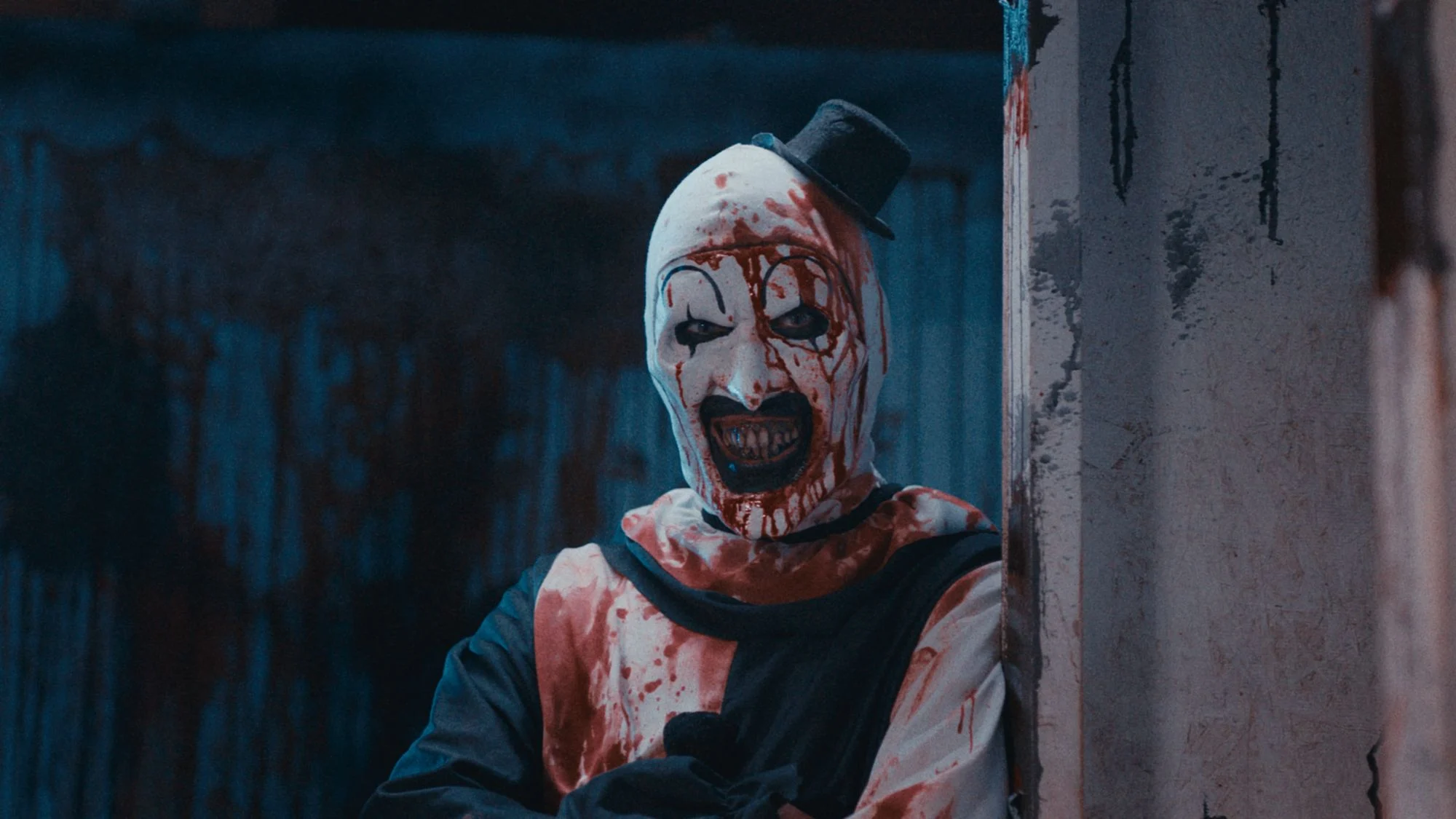 Cena do filme Terrifier: O Início mostra o palhaço ensanguentado encostado em uma parede